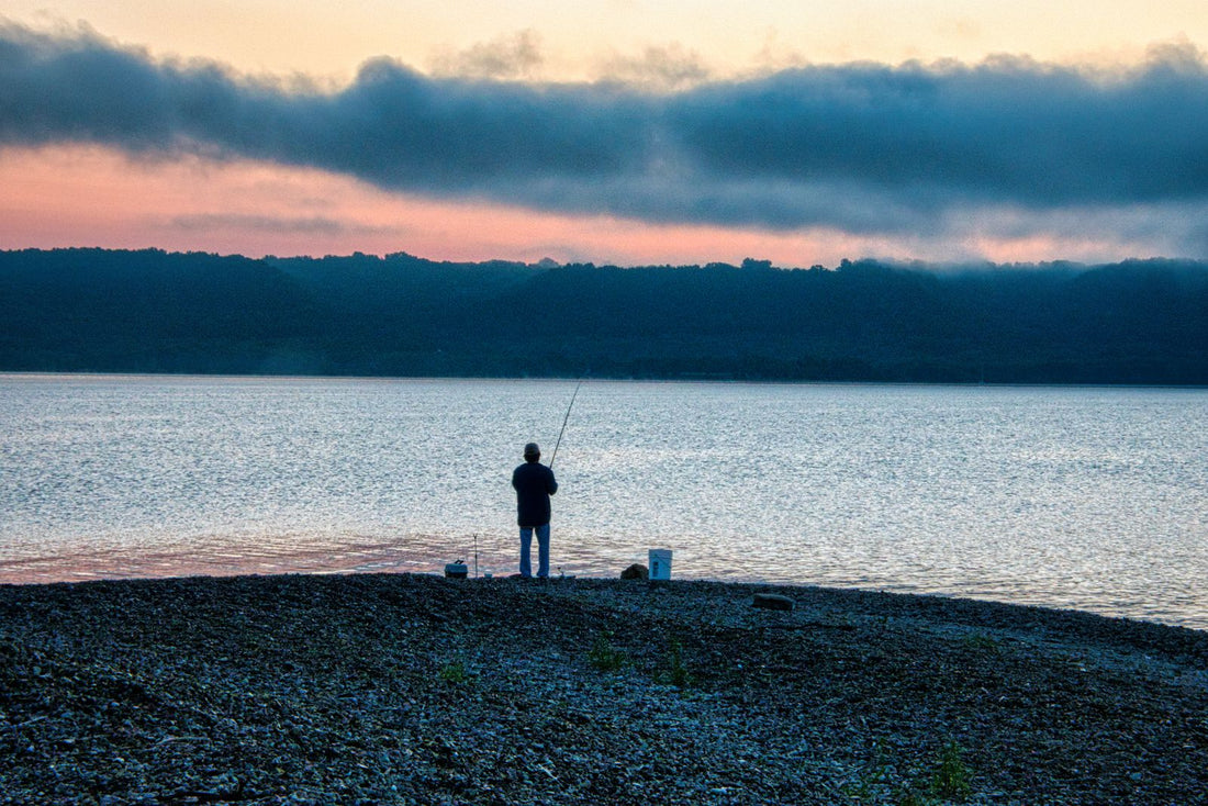 One angler fishing from a stony lakeshore beneath a pink and blue dawn sky