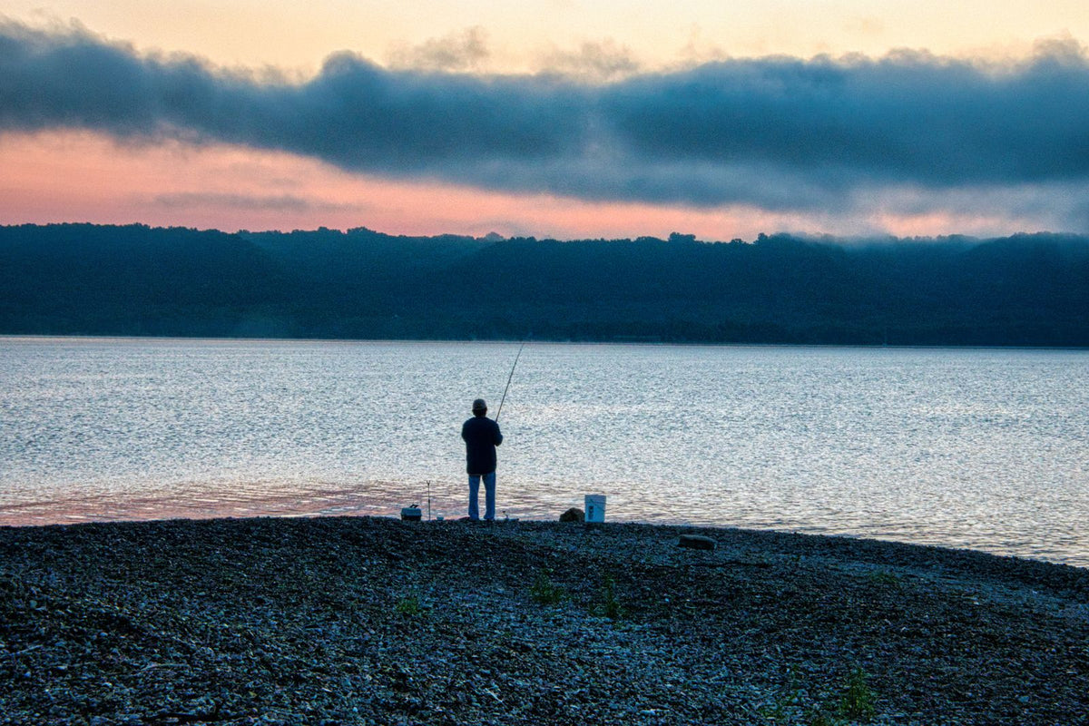 One angler fishing from a stony lakeshore beneath a pink and blue dawn sky