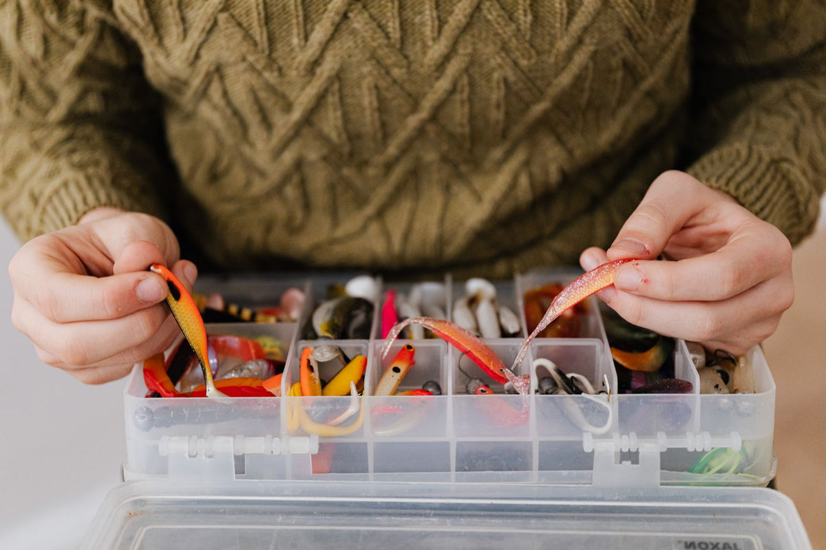 Hands comparing two colorful soft plastic baits above an open compartmented tackle box